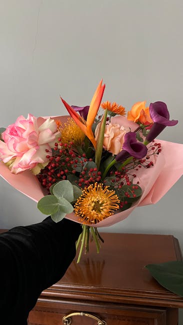 A vibrant floral bouquet held up against a plain light-colored wall, featuring a mix of fresh flowers including purple calla lilies, orange tulips, pink roses, a striking orange bird of paradise, yell