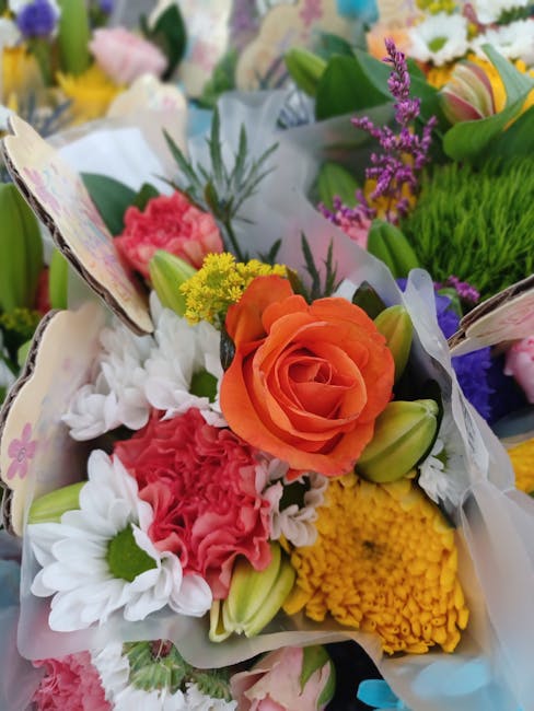 A vibrant floral bouquet featuring a large orange rose at the center, surrounded by white daisies, pink carnations, yellow chrysanthemums, and sprigs of purple lavender. The flowers are arranged in a 